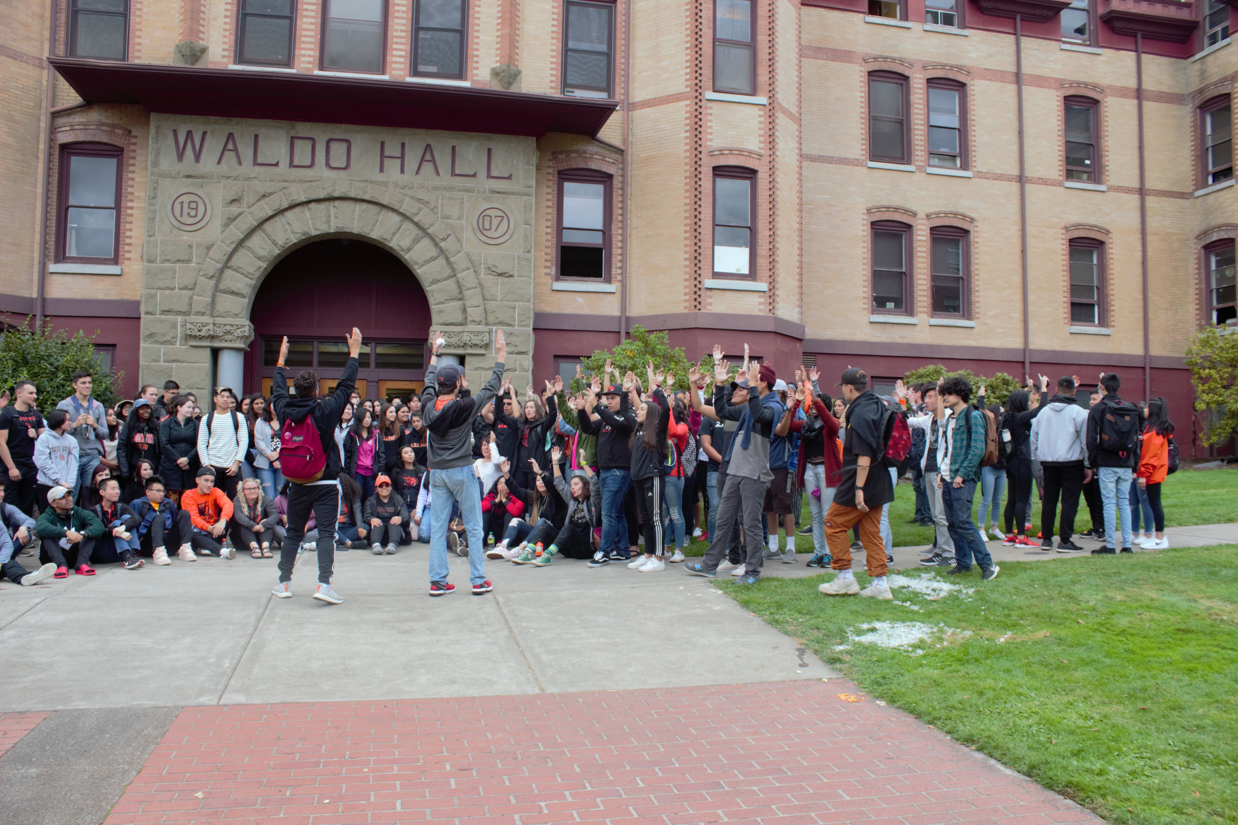 Group of students standing outside of campus building with arms raised