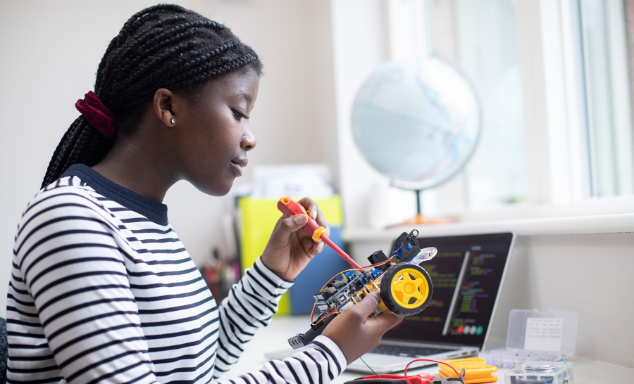 Black female student tinkering with robot seated at desk next to laptop