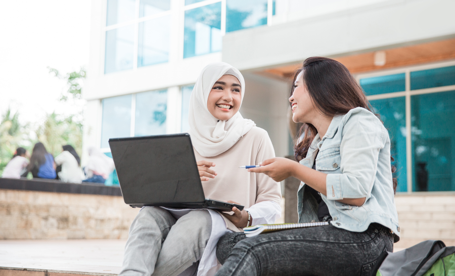 Two female students, one of whom is wearing a hijab, sit outside and work on a laptop