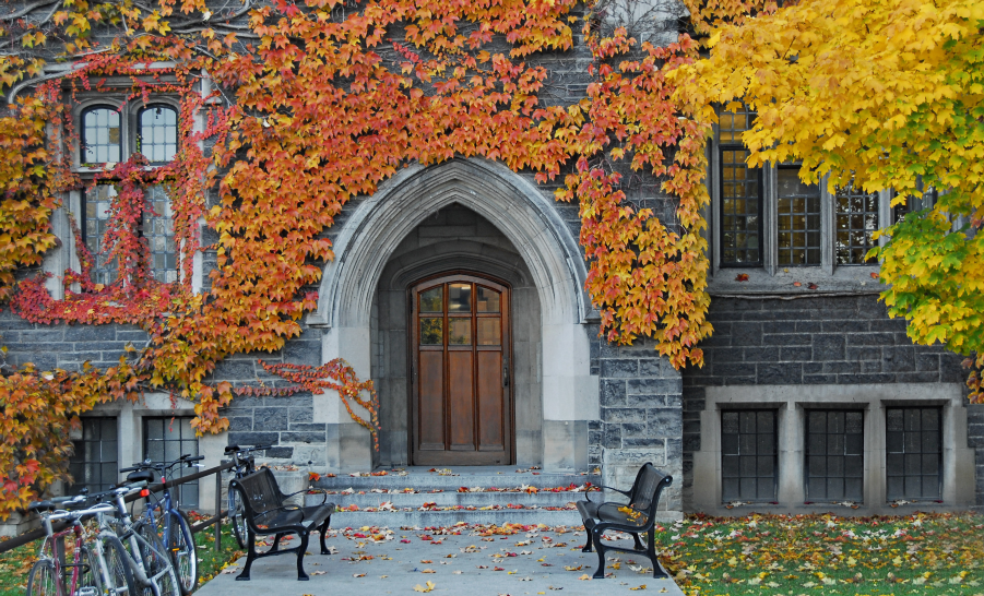 Door to stone campus building covered in fall foliage flanked by wooden benches and a bicycle rack
