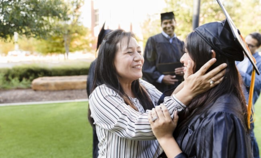 Mother holding daughter's face and smiling outside on daughter's graduation day while other graduates look on